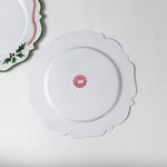 reverse side of A white tole metal christmas charger with a pink ribbon and holly motifs around the edges against a white background.  