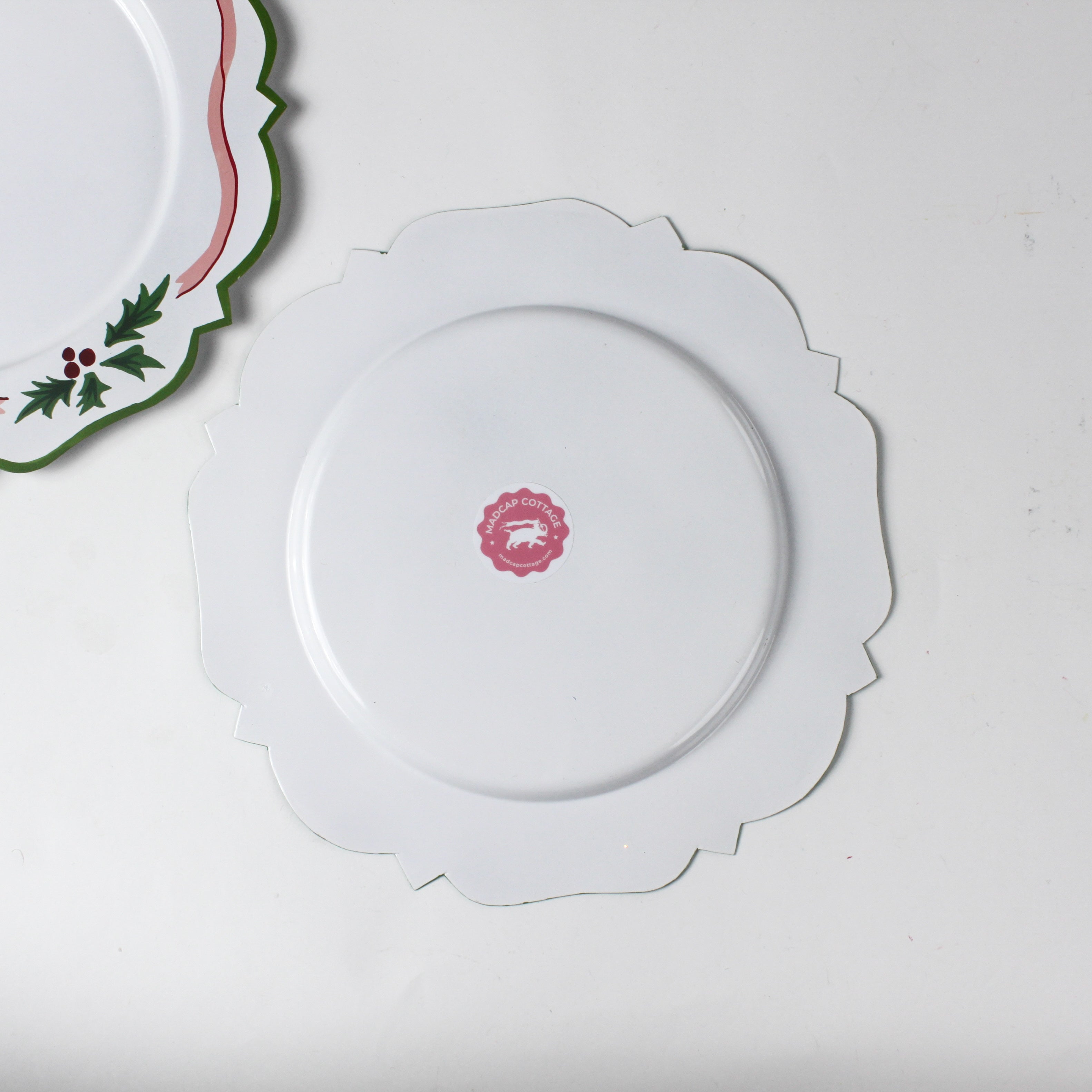 reverse side of A white tole metal christmas charger with a pink ribbon and holly motifs around the edges against a white background.  