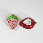 a pair of small ceramic strawberry shaped bowls, one red, one pink on a white background.