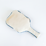 The back side of a pickleball paddle shaped pillow featuring a soft cream fabric surface with blue trim edges.