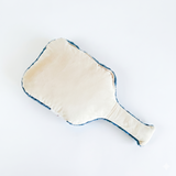 The back side of a pickleball paddle shaped pillow featuring a soft cream fabric surface with blue trim edges.