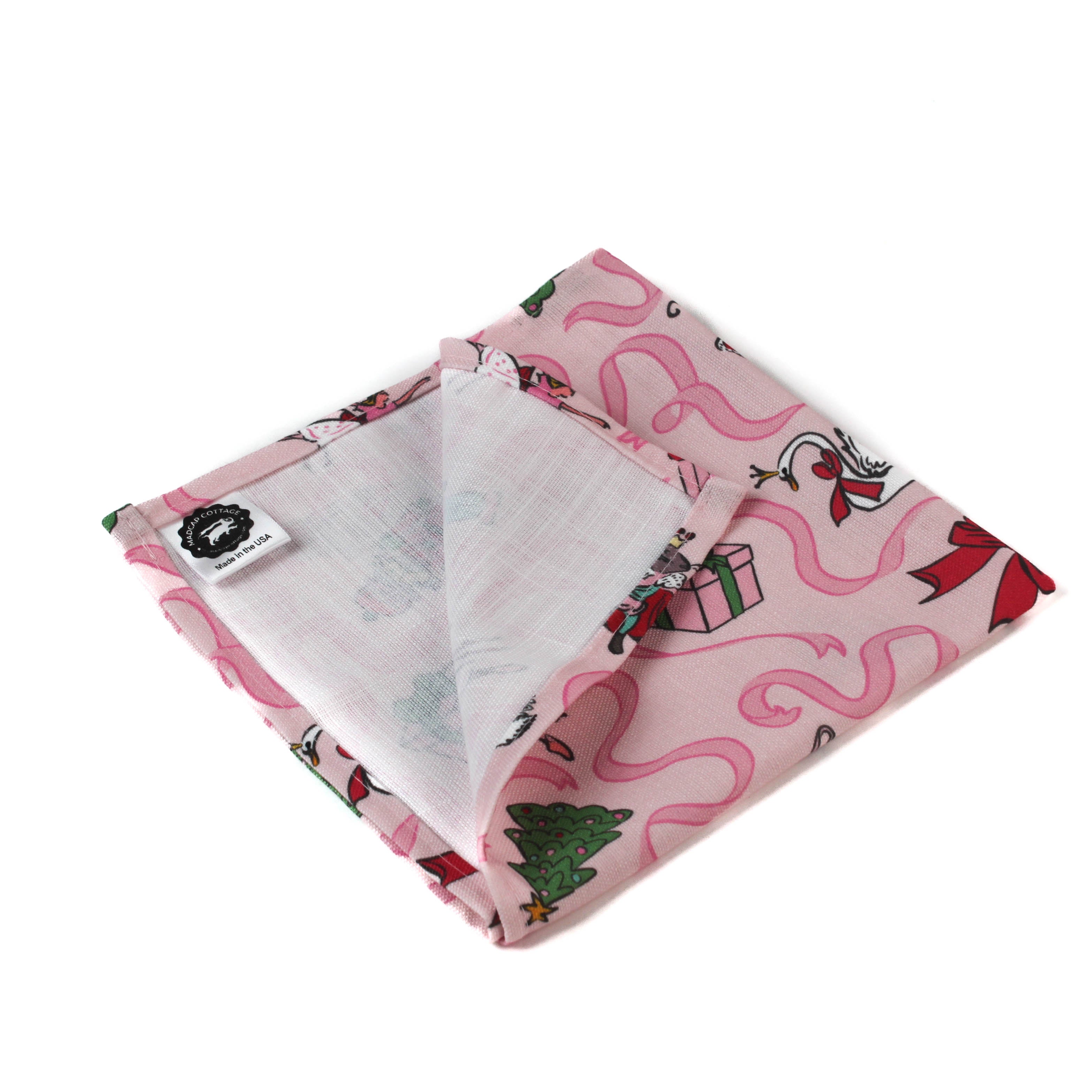 a folded Nutcracker pink christmas napkin featuring motifs of ballerinas, ribbons, christmas trees and nutcrackers on a white background. 