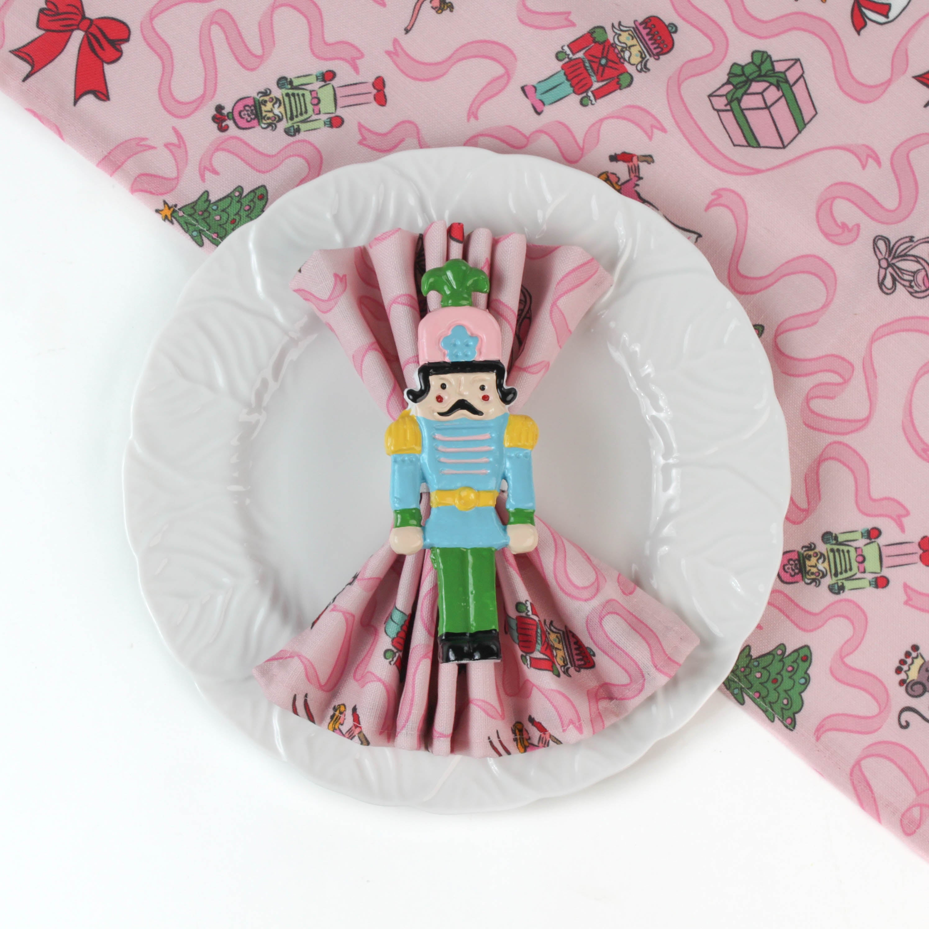 a white plate with a Nutcracker pink christmas napkinsfeaturing motifs of ballerinas, ribbons, christmas trees and nutcrackers on top of a matching christmas table runner on a white background. 
