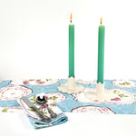 Green and blue ricrac stripe taper candles lit on a spring table setting with floral table linens and candle holders.