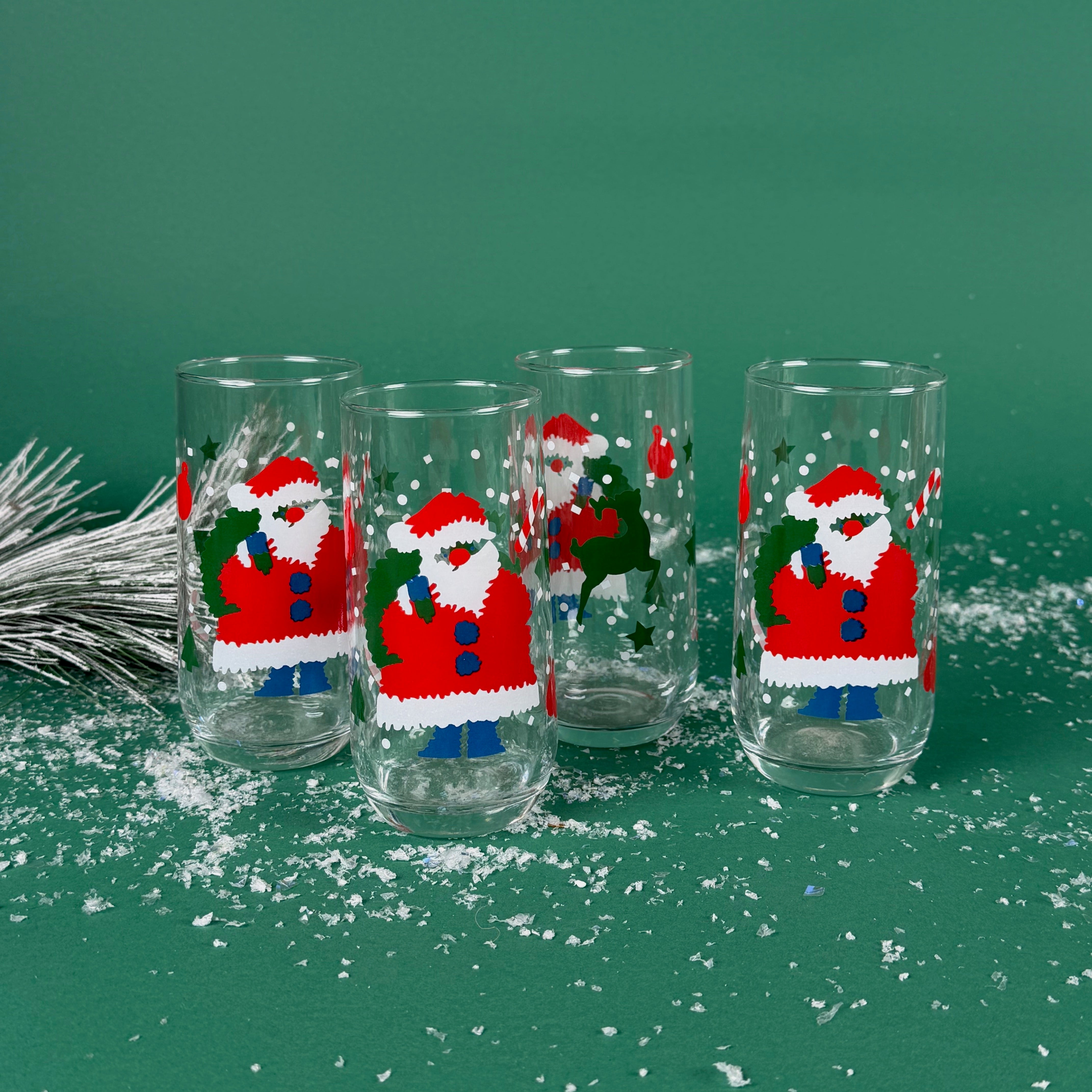 Set of tall clear drinking glasses with colorful Santa Claus designs on a snowy green background
