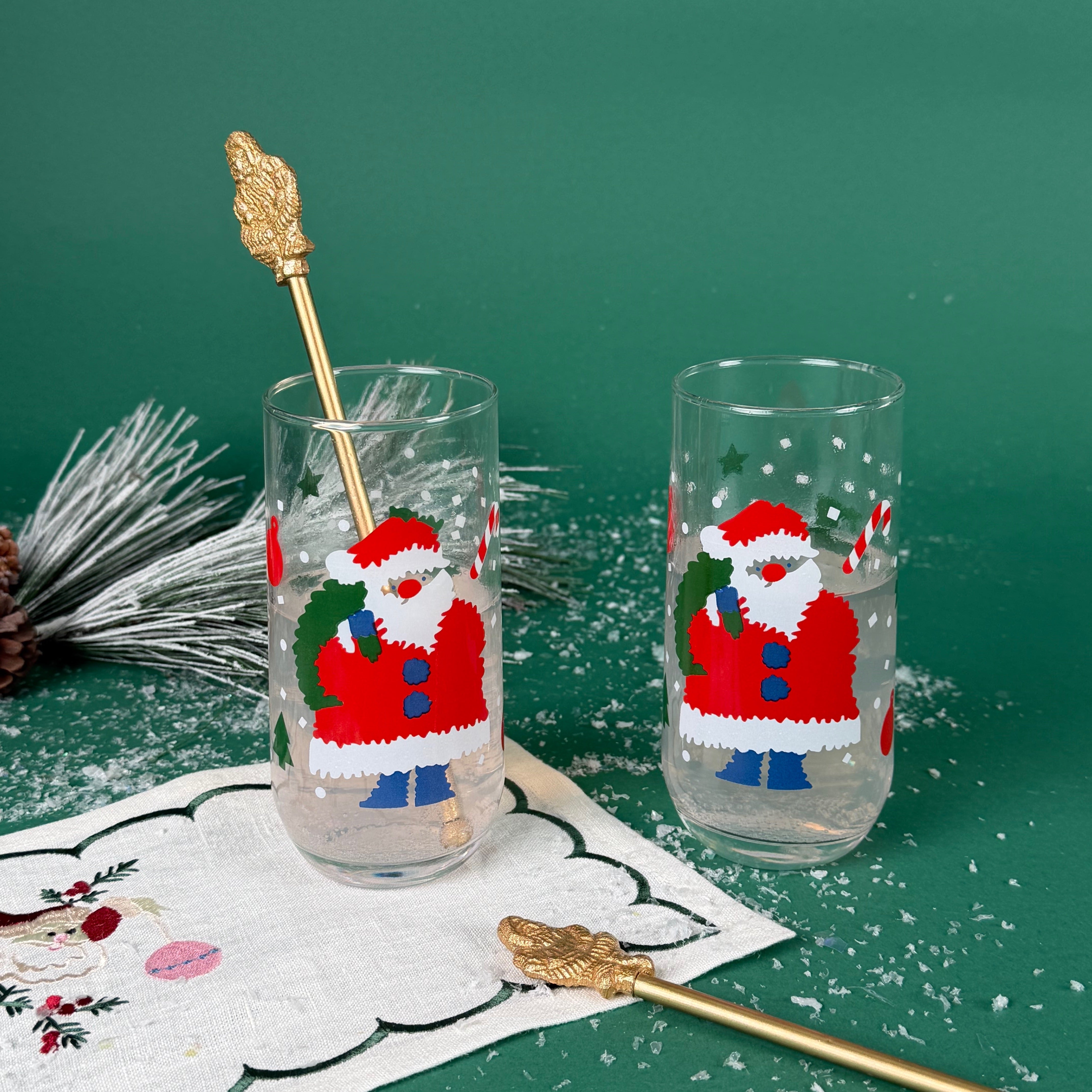 Two clear drinking glasses with festive Santa Claus designs, holiday stir sticks, and faux snow on a green background