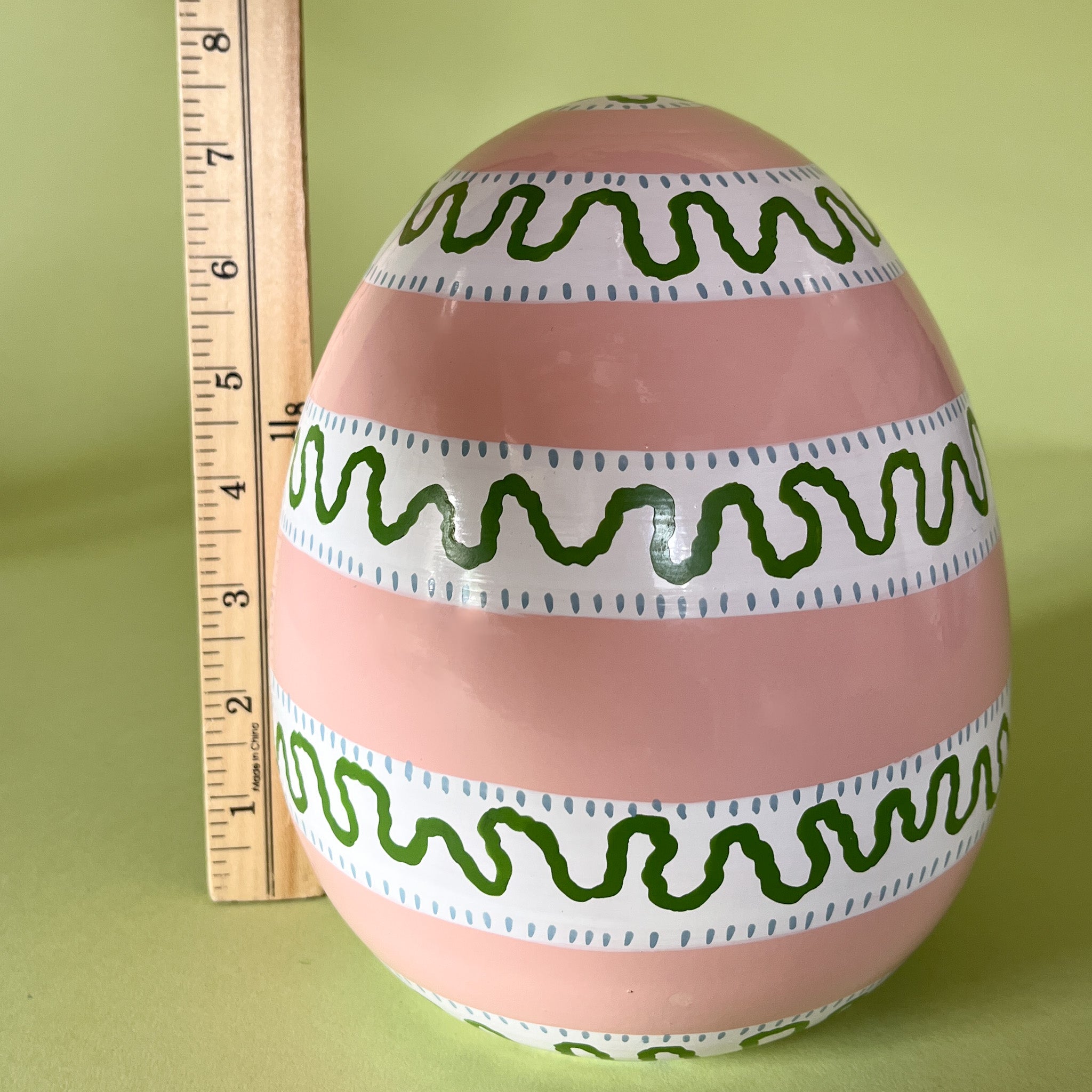 a striped pink large decorative easter egg in tole metal and a ruler in the background to illustrate scale