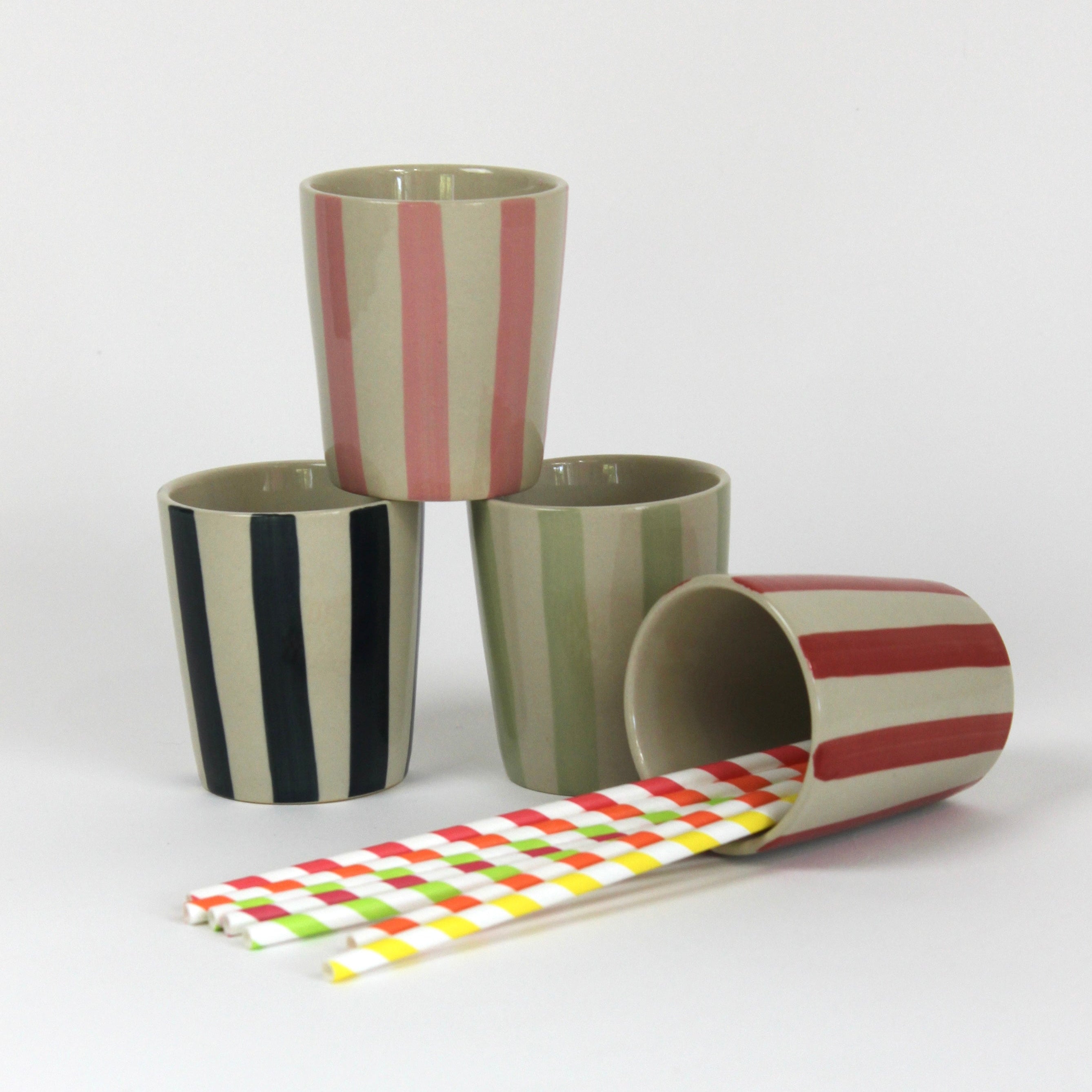 A set of four striped ceramic cups in blue, green, pink and red with straws in a white background