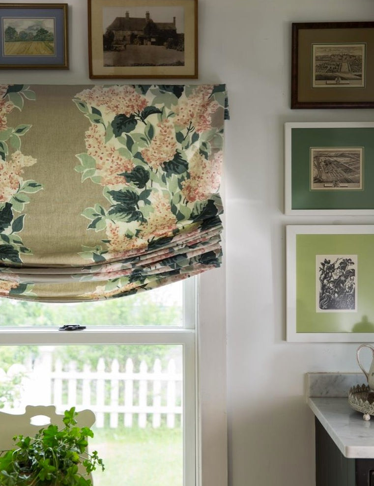 Floral patterned Roman shade in soft green and cream tones styled in a cozy room with framed wall art and natural light.