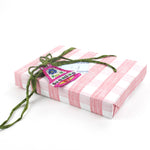 Rectangular gift wrapped in pink plaid wrapping paper, decorated with green ribbon and bell ornament.