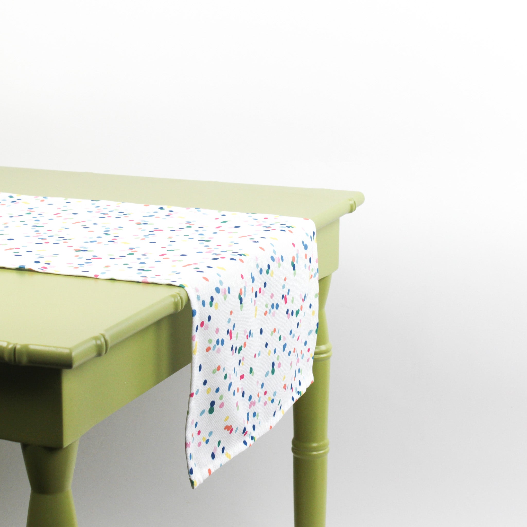 Side view of a white table runner with multicolor confetti dots draped over a green table.