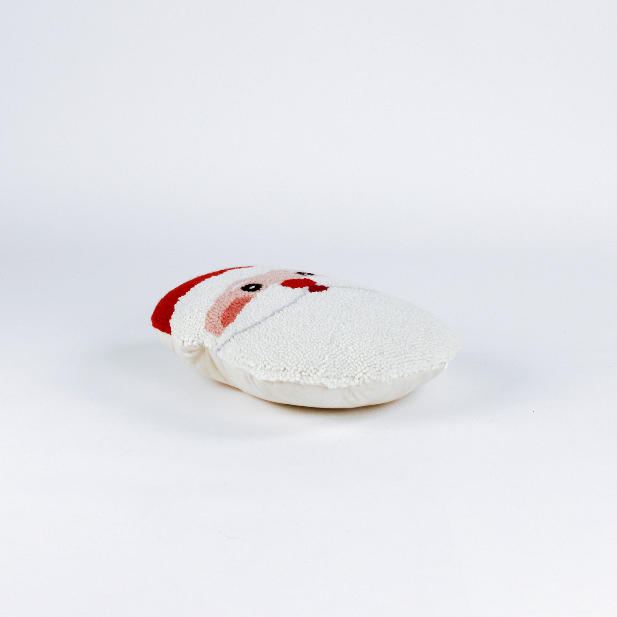 side of a Santa-shaped christmas pillow with a hooked texture, featuring Santa’s face with rosy cheeks, a red nose, and a bright red hat with a white pom-pom.
