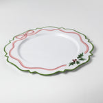 A white tole metal christmas charger with a pink ribbon and holly motifs around the edges against a white background