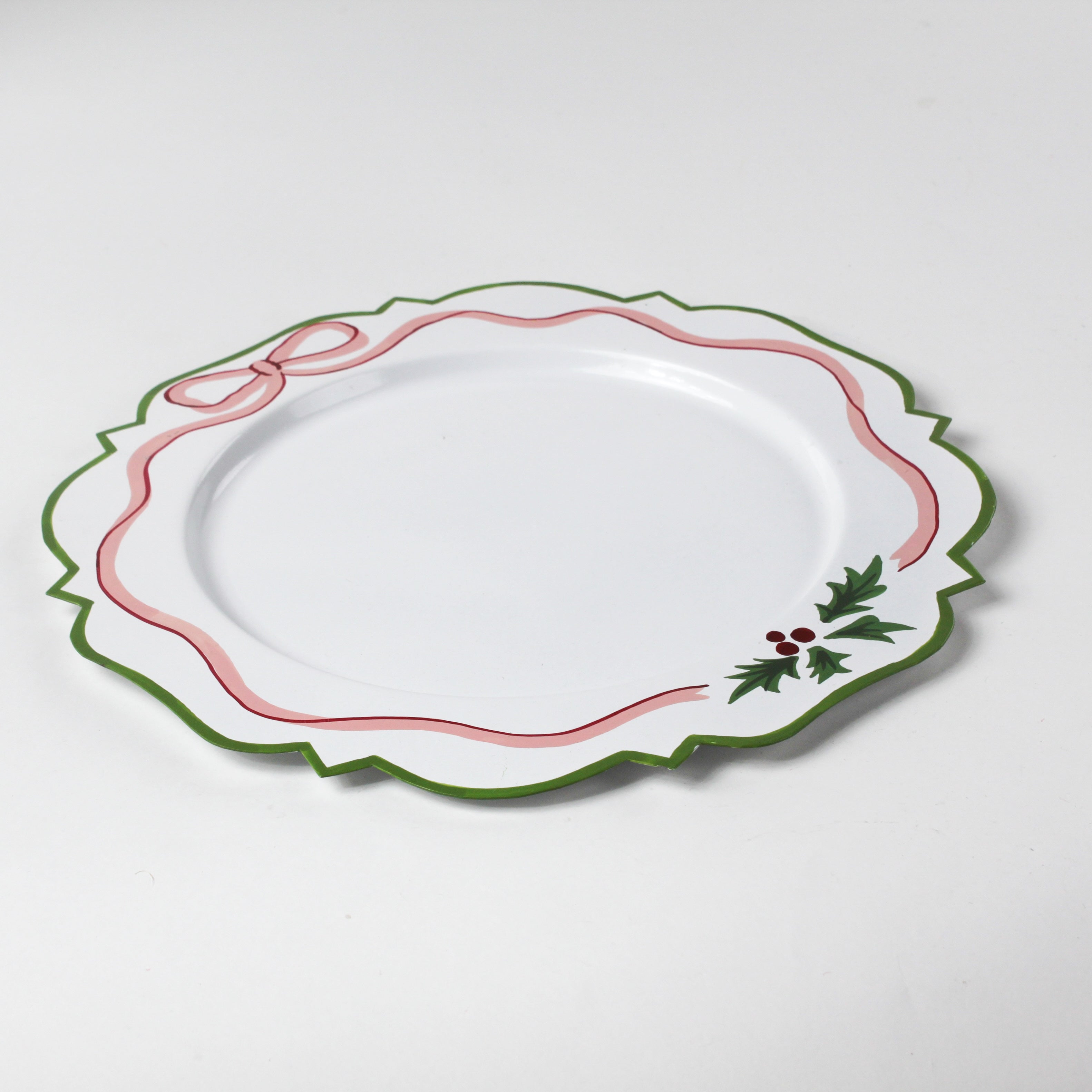 A white tole metal christmas charger with a pink ribbon and holly motifs around the edges against a white background
