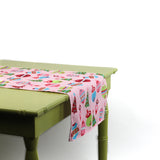 Close-up angled view of a pink table runner with colorful Christmas ornament prints draped over a green table.