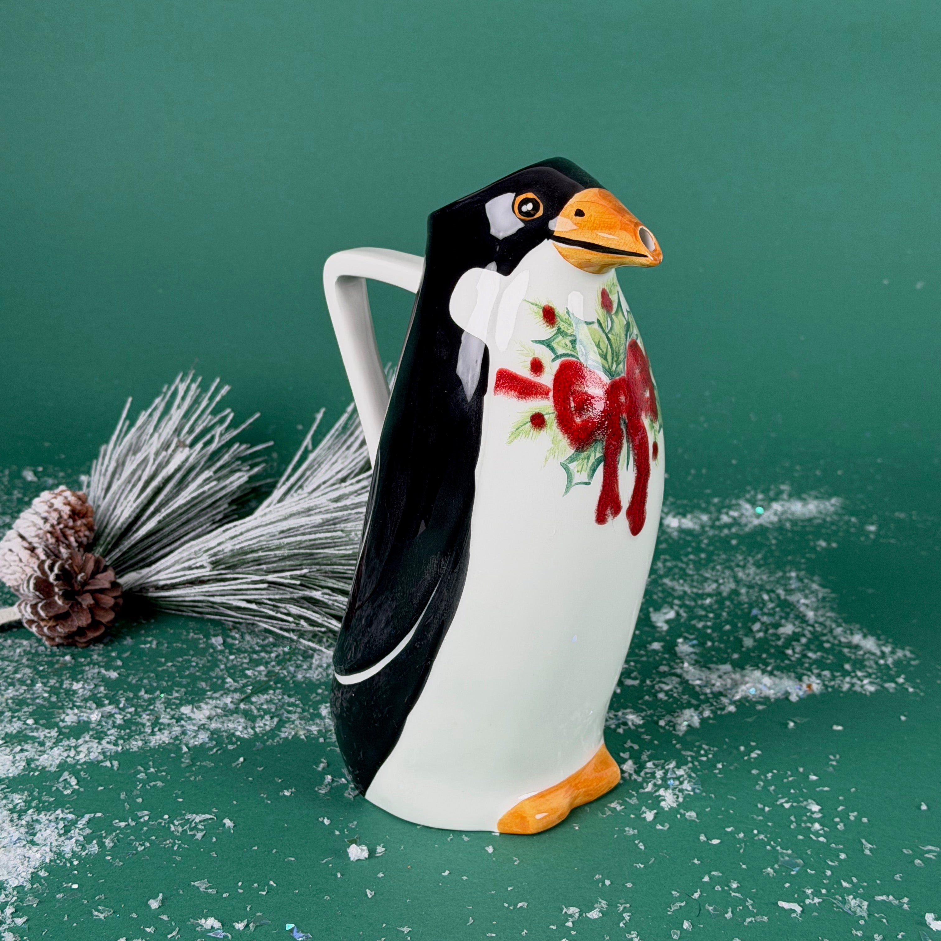 Ceramic penguin-shaped holiday pitcher with a red bow and holly design on a snowy green background