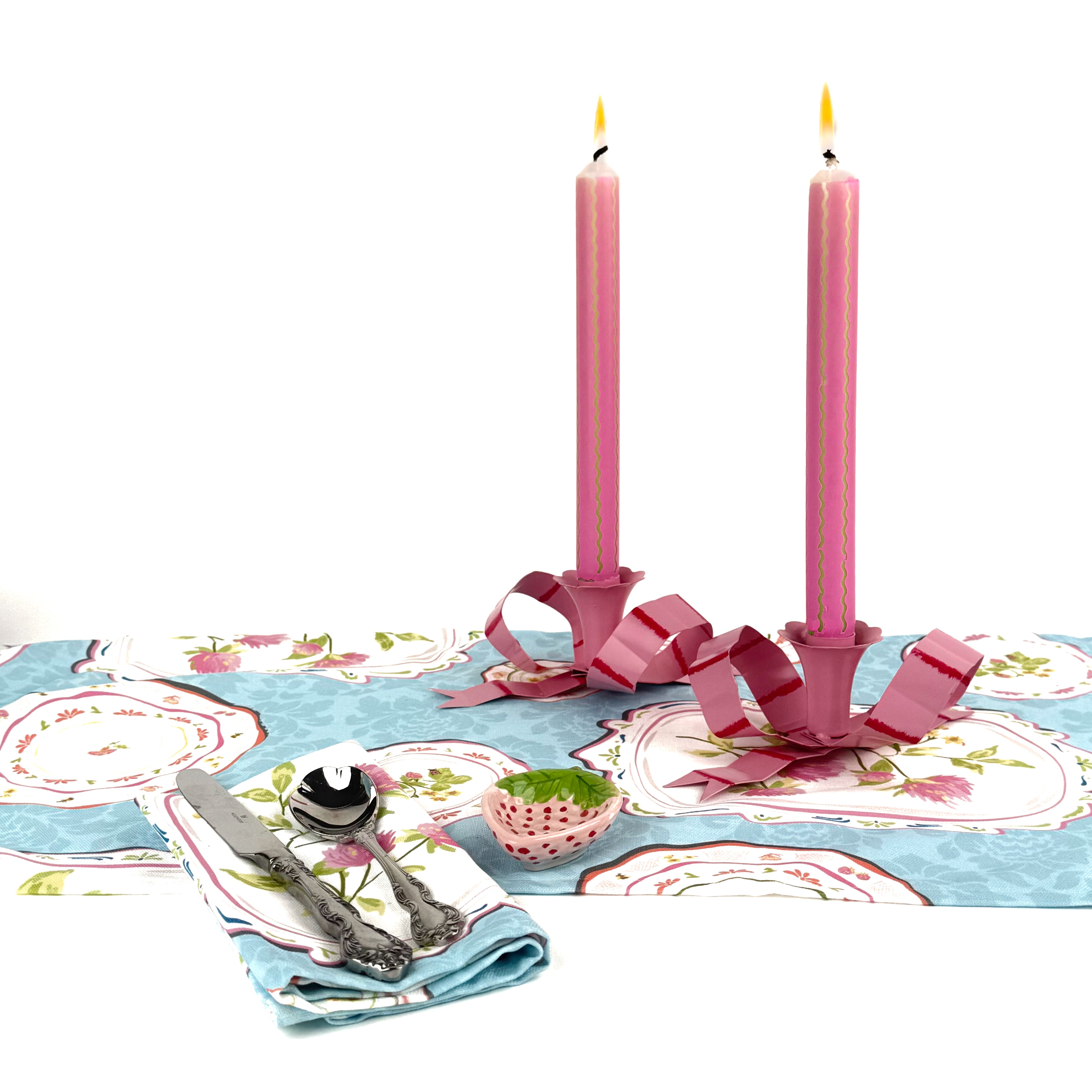 Pink taper candles with green squiggle stripes lit on a spring tablescape with floral table linens, pink candle holders, and silverware.

