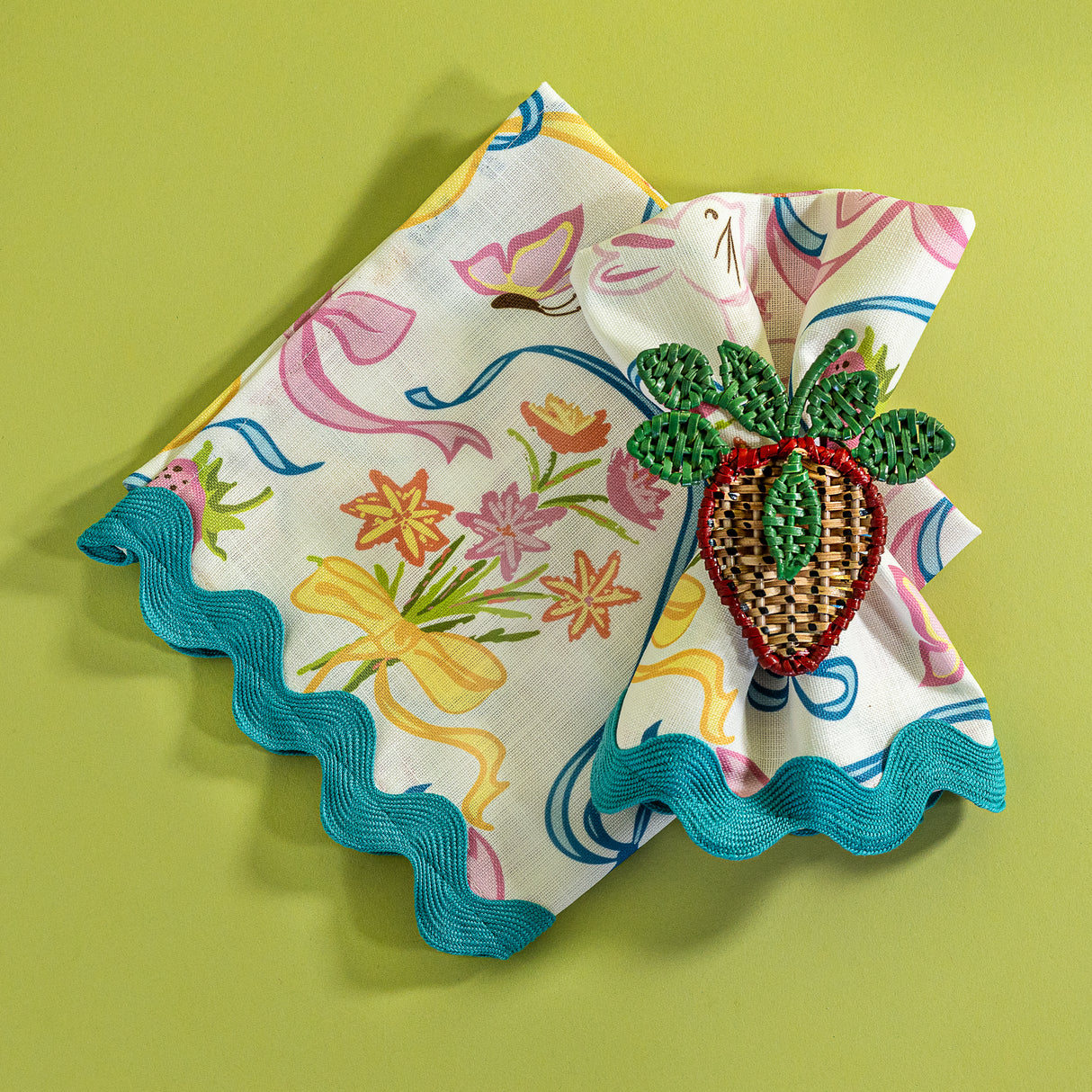 A Bunnies and bow print cloth dinner napkins in multicolor with a strawberry napkin ring against a light green background.