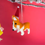 Corgi Christmas ornament with a Santa hat hanging from a tree branch against a red background