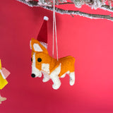 Corgi Christmas ornament with a Santa hat hanging from a tree branch against a red background