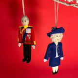Two Christmas British Royal Family ornaments of King Charles and Queen Camilla anging from a branch against a red background.