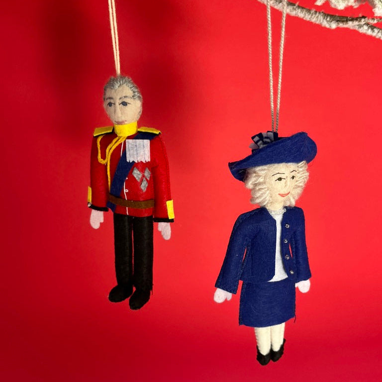 Two Christmas British Royal Family ornaments of King Charles and Queen Camilla anging from a branch against a red background.