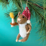 A felt mouse ornament wearing a red patterned nightcap holds a small white candle on a brown base, hanging from a pine branch against a teal background.

