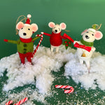 Three festive felt mouse ornaments dressed in holiday outfits stand on fake snow, holding candy canes, with pine branches and candy decorations around them.