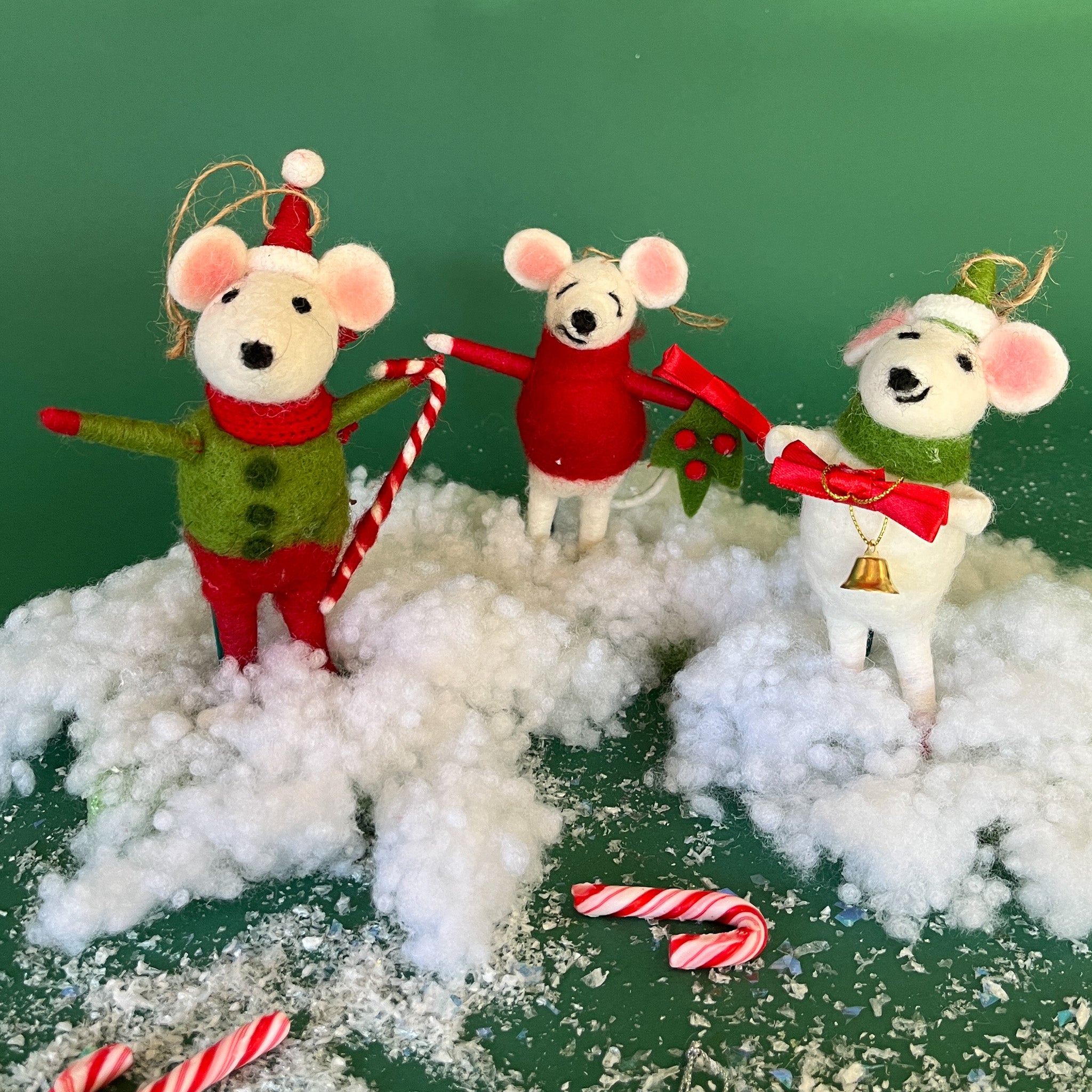 Three festive felt mouse ornaments dressed in holiday outfits stand on fake snow, holding candy canes, with pine branches and candy decorations around them.