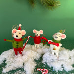 Three festive felt mouse ornaments dressed in holiday outfits stand on fake snow, holding candy canes, with pine branches and candy decorations around them.