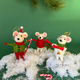 Three festive felt mouse ornaments dressed in holiday outfits stand on fake snow, holding candy canes, with pine branches and candy decorations around them.