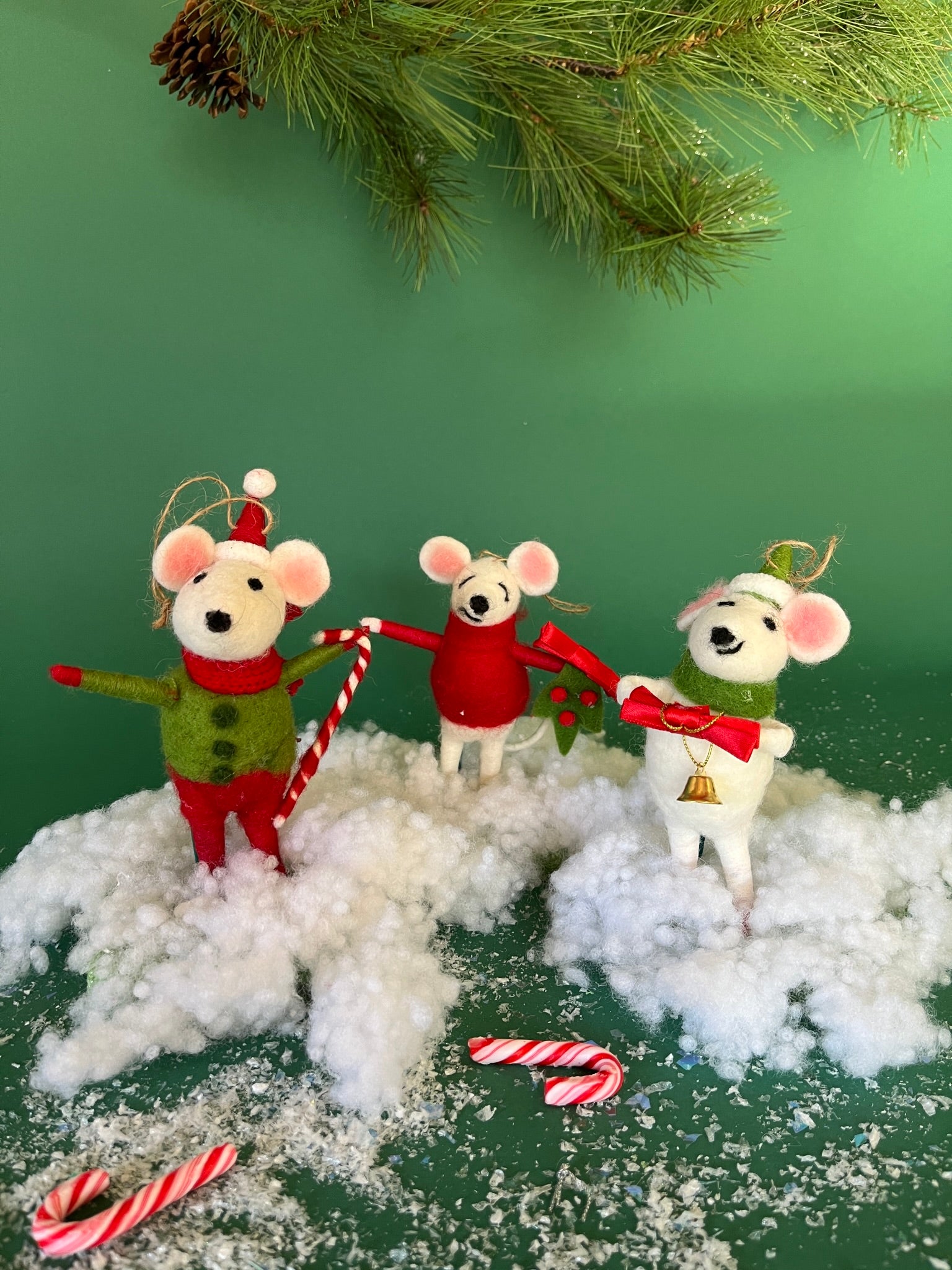 Three festive felt mouse ornaments dressed in holiday outfits stand on fake snow, holding candy canes, with pine branches and candy decorations around them.