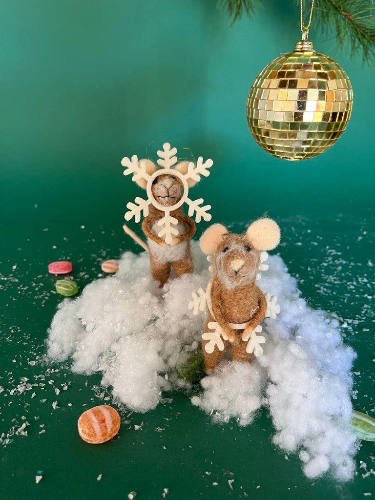 Two felt mouse ornaments dressed as snowflakes stand on fake snow beneath a gold disco ball, surrounded by scattered candy on a green background.