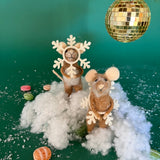 Two felt mouse ornaments dressed as snowflakes stand on fake snow beneath a gold disco ball, surrounded by scattered candy on a green background.