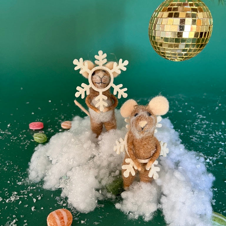 Two felt mouse ornaments dressed as snowflakes stand on fake snow beneath a gold disco ball, surrounded by scattered candy on a green background.