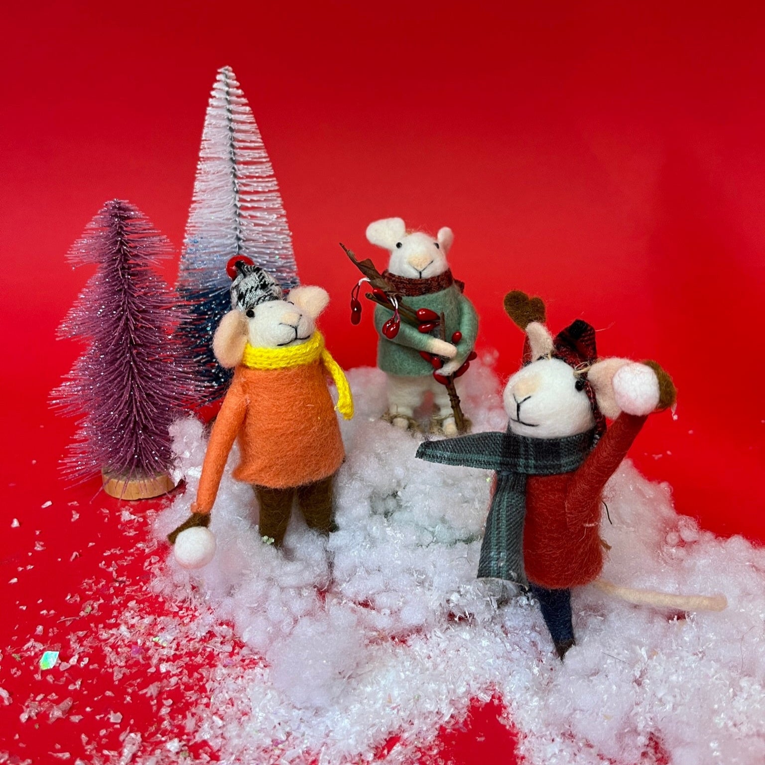 Three felt mouse ornaments dressed for winter—one in a red coat and plaid hat tossing a snowball, one in an orange coat with a yellow scarf holding a snowball, and one in a green sweater holding red berries. Playing in faux snow among mini Christmas trees.