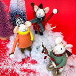 close up Three felt mouse ornaments dressed for winter—one in a red coat and plaid hat tossing a snowball, one in an orange coat with a yellow scarf holding a snowball, and one in a green sweater holding red berries. Playing in faux snow among mini Christmas trees.