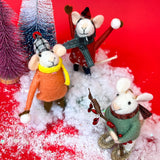 close up Three felt mouse ornaments dressed for winter—one in a red coat and plaid hat tossing a snowball, one in an orange coat with a yellow scarf holding a snowball, and one in a green sweater holding red berries. Playing in faux snow among mini Christmas trees.