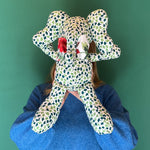 Green and white patterned stuffed elephant toy with a red bow hiding behind.