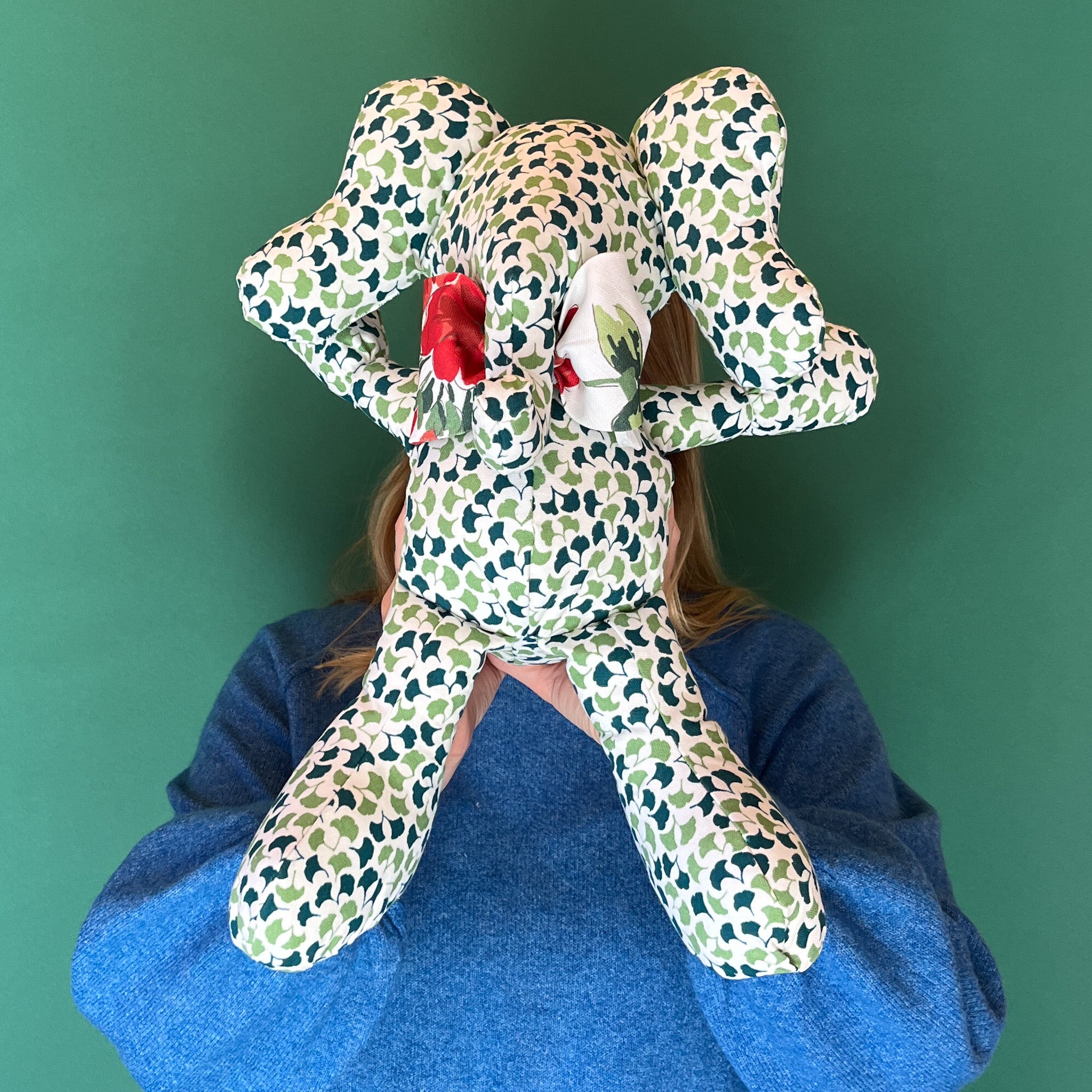 Green and white patterned stuffed elephant toy with a red bow hiding behind.