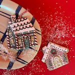 Two gingerbread house Christmas ornaments, one pink and one brown with candy cane details, shown with red and white hanging strings—displayed on a festive red background with faux snow and a striped plate.
