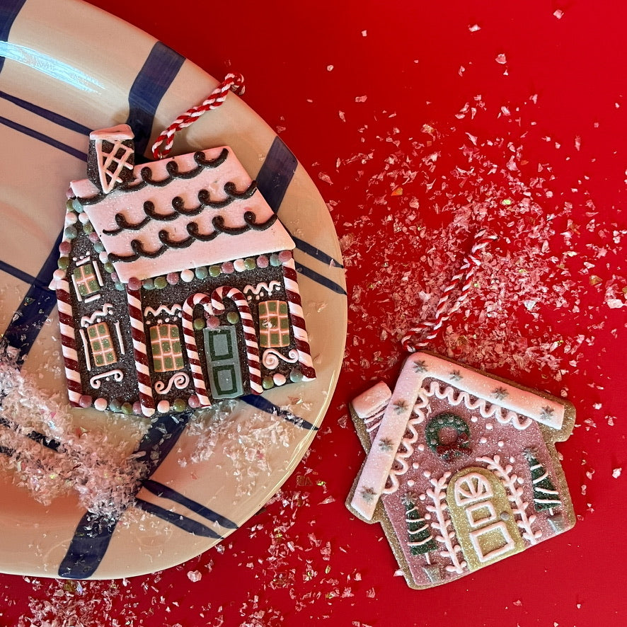 Two gingerbread house Christmas ornaments, one pink and one brown with candy cane details, shown with red and white hanging strings—displayed on a festive red background with faux snow and a striped plate.