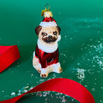 Pug dog Christmas ornament in a Santa hat and scarf on a green background with red ribbon and gift box.