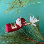 a santa riding a red champagne bottle ornament hanging in a branch against a green background