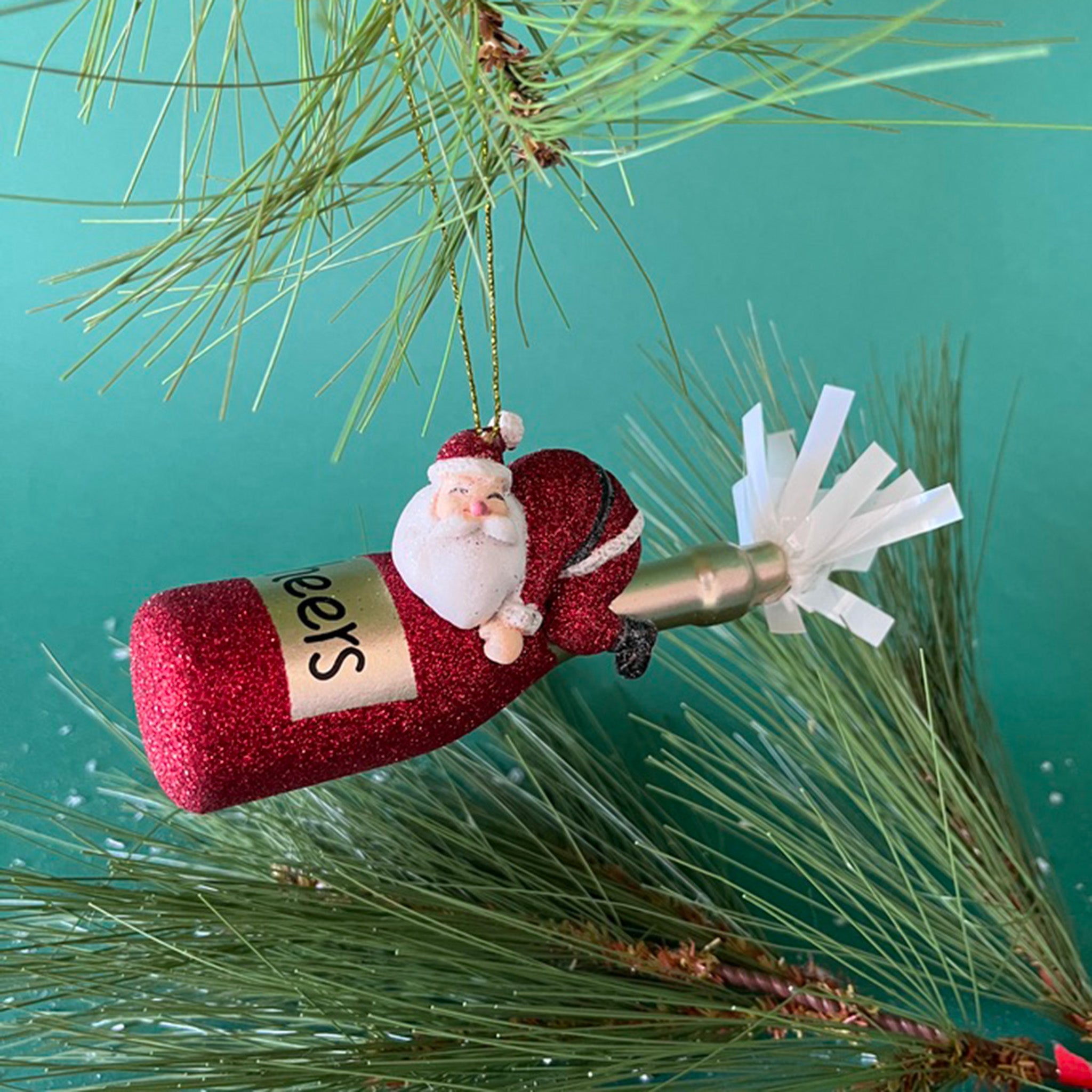 a santa riding a red champagne bottle ornament hanging in a branch against a green background
