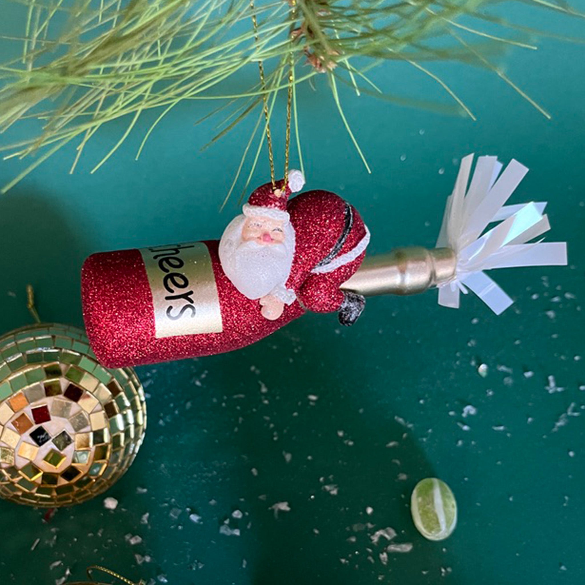 a santa riding a red champagne bottle ornament hanging in a branch against a green background
