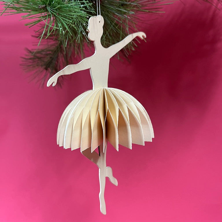 Paper ballerina ornament hanging on a Christmas tree with a pink background