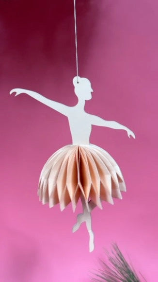 Decorative paper ballerina christmas ornament hanging against a pink background
