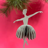 3D ballerina christmas ornament hanging from a Christmas tree branch against a red background