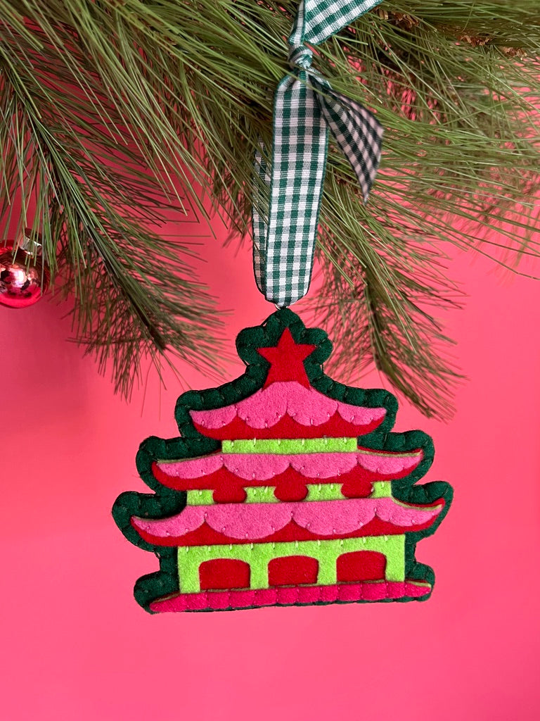 Decorative Christmas chinoiserie ornament shaped like a pagoda hanging on a tree branch with a pink background.